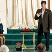 Marty G smiling and holding up a single playing card while performing magic trick on stage, a young girl seated beside him watching eagerly.