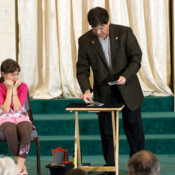 Marty G wearing dark suit jacket performing magic trick on stage with young girl seated beside him watching him expectantly.