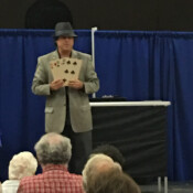 Marty G wearing fedora performing magic trick involving three oversize playing cards: six of spades, six of clubs, and six of hearts.