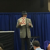 Marty G wearing fedora performing magic trick involving three oversize playing cards: six of spades, six of clubs, eight of hearts.