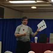 Marty G smiles at seated audience while performing magic trick involving three oversize playing cards.