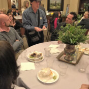 Marty G wearing fedora performs magic trick with playing cards for several guests seated at a formal dinner table at a reception with many other guests.