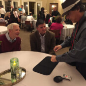 Two men seated at a wedding reception dining table eagerly watch Marty G one perform magic trick involving sleight of hand.